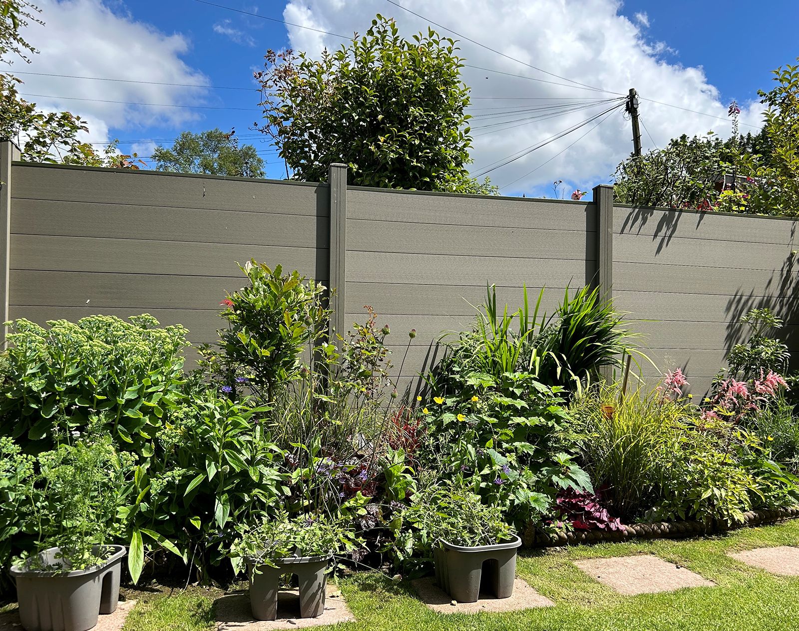 Light brown composite fencing panels providing a modern garden backdrop behind a vibrant flower bed filled with mixed plants and greenery.
