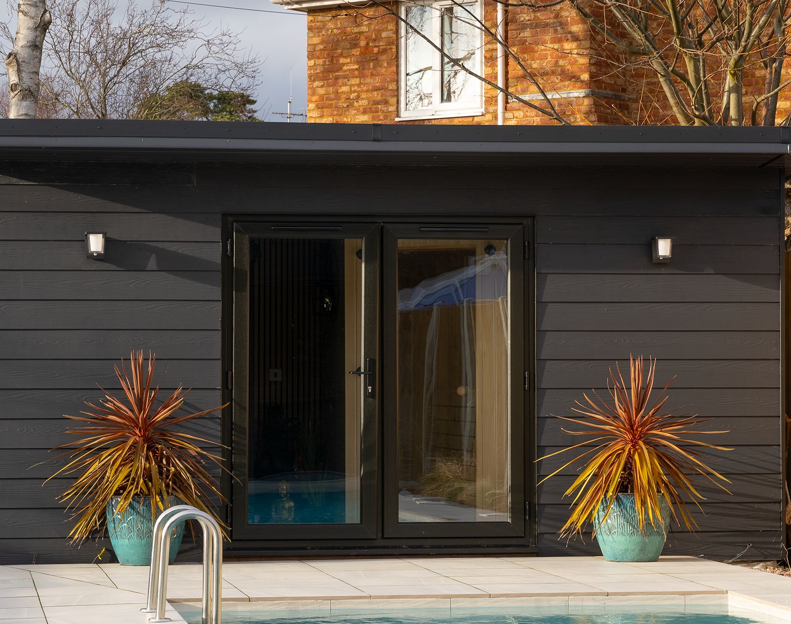 Modern garden room with dark fibre cement cladding and black-framed double doors, set beside a swimming pool with tropical plants in turquoise pots.