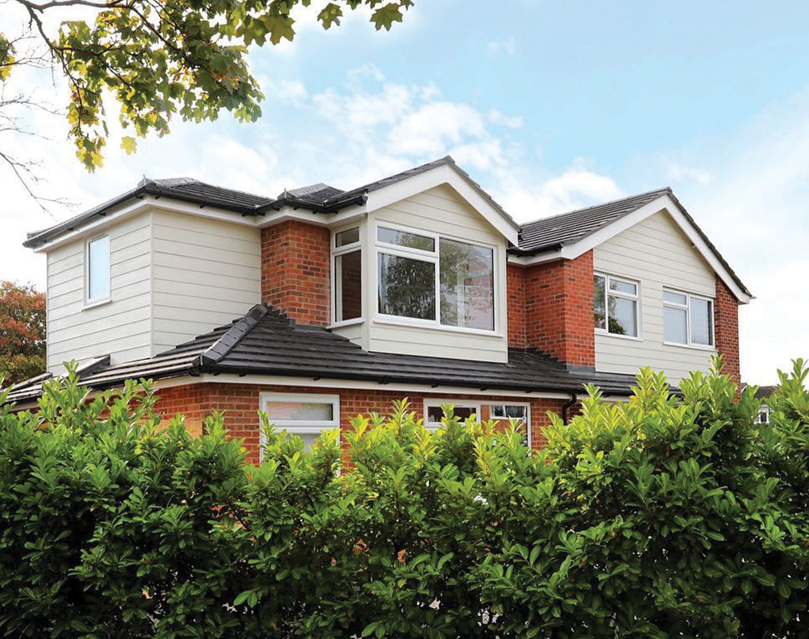 Two-storey house with cream fibre cement cladding on the upper floor and red brickwork on the ground floor, surrounded by greenery.