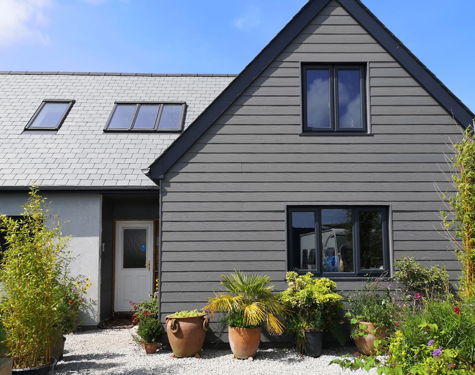Modern home exterior featuring light grey fibre cement cladding and slate roof, with potted plants and gravel landscaping in the front garden.