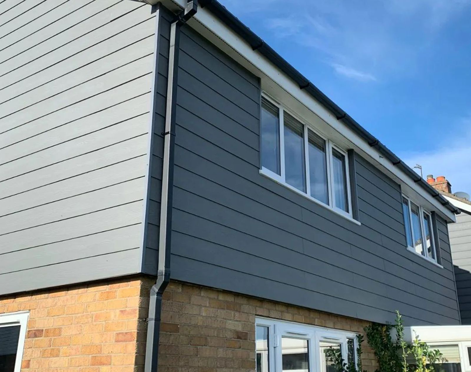 Grey fibre cement cladding fitted to the upper level of a modern two-storey house with brickwork on the ground floor and white framed windows.