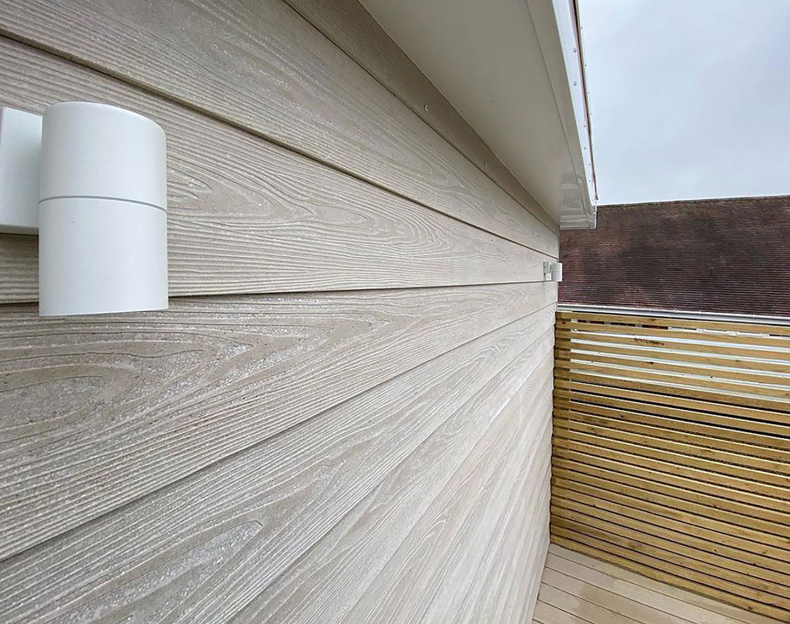 Close-up of beige woodgrain-effect fibre cement cladding on a balcony wall with modern white light fitting and slatted timber privacy screen.