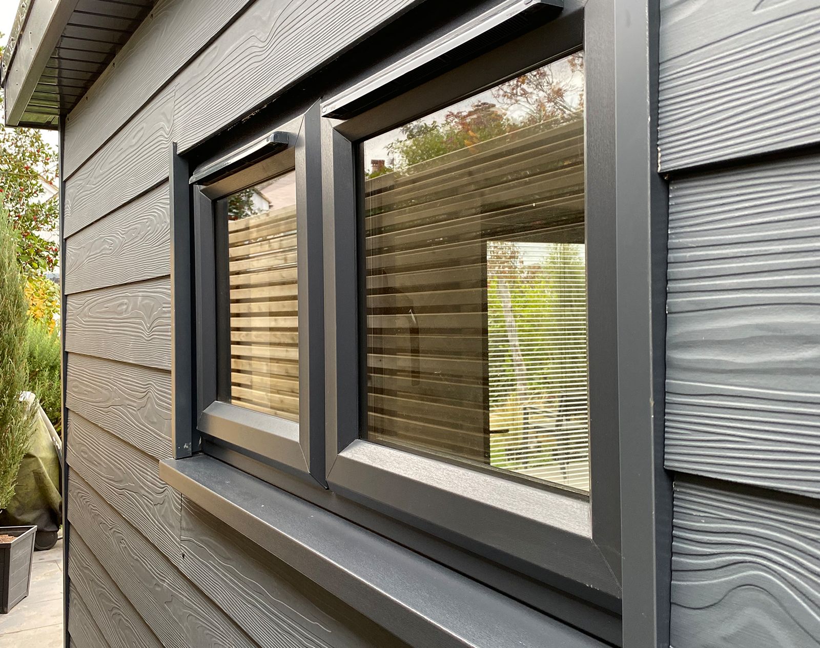 Dark grey fibre cement cladding with woodgrain texture surrounding modern double-glazed windows with anthracite frames.