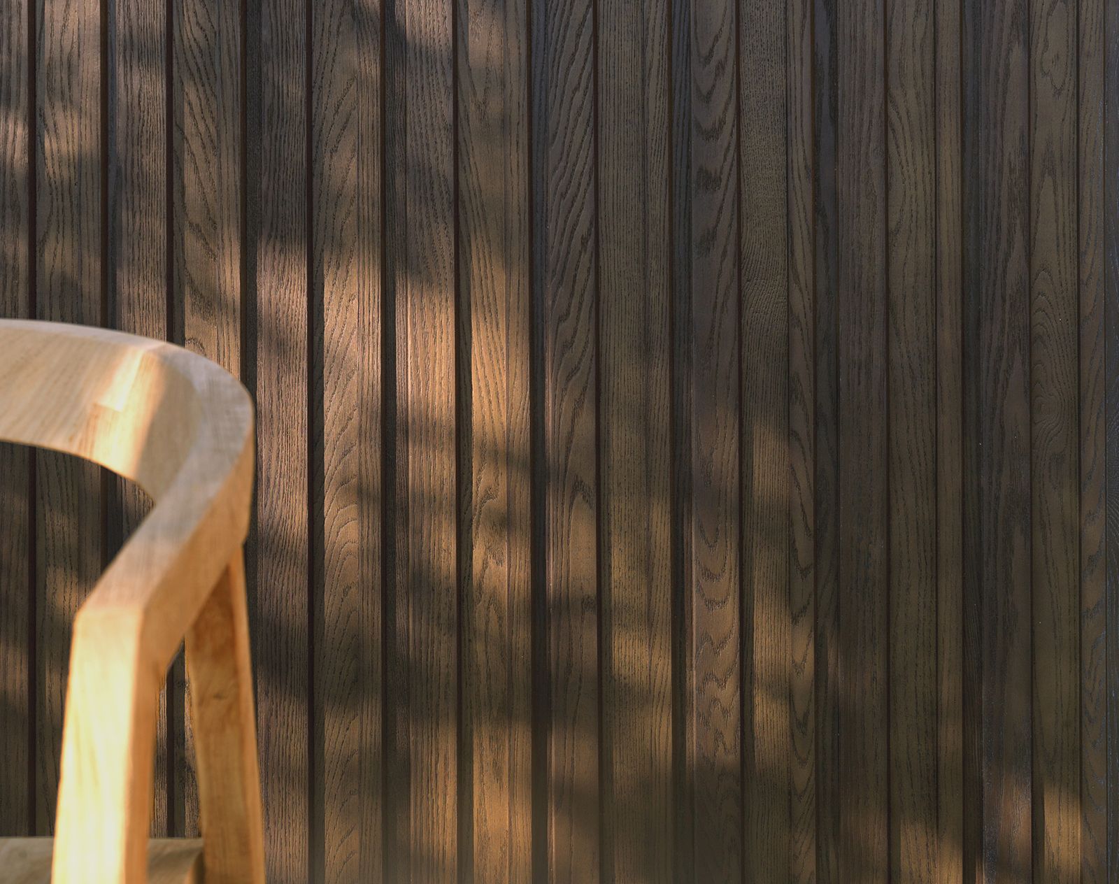 Shadow-lit vertical Millboard cladding in dark woodgrain finish with part of a curved timber chair in the foreground.