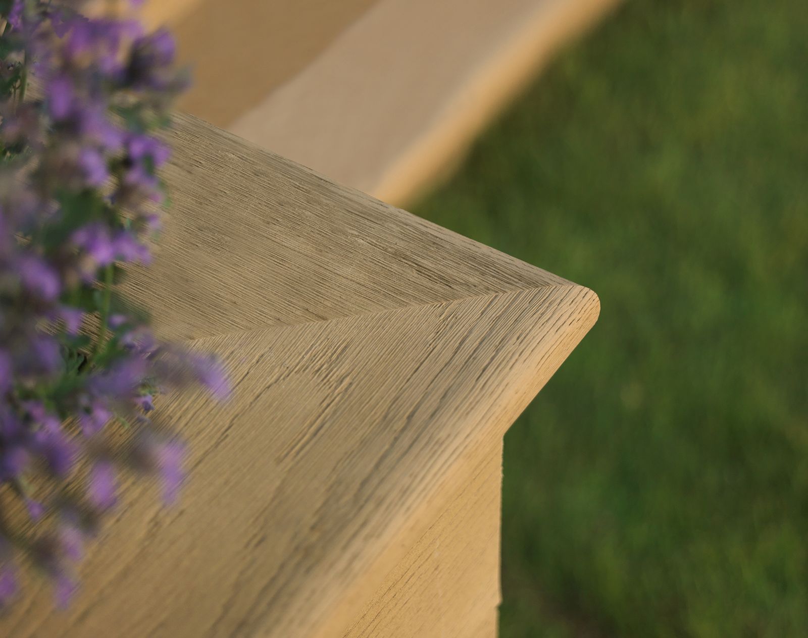 Close-up of a chamfered corner on golden Millboard decking, highlighting wood grain detail next to lawn and lavender flowers.