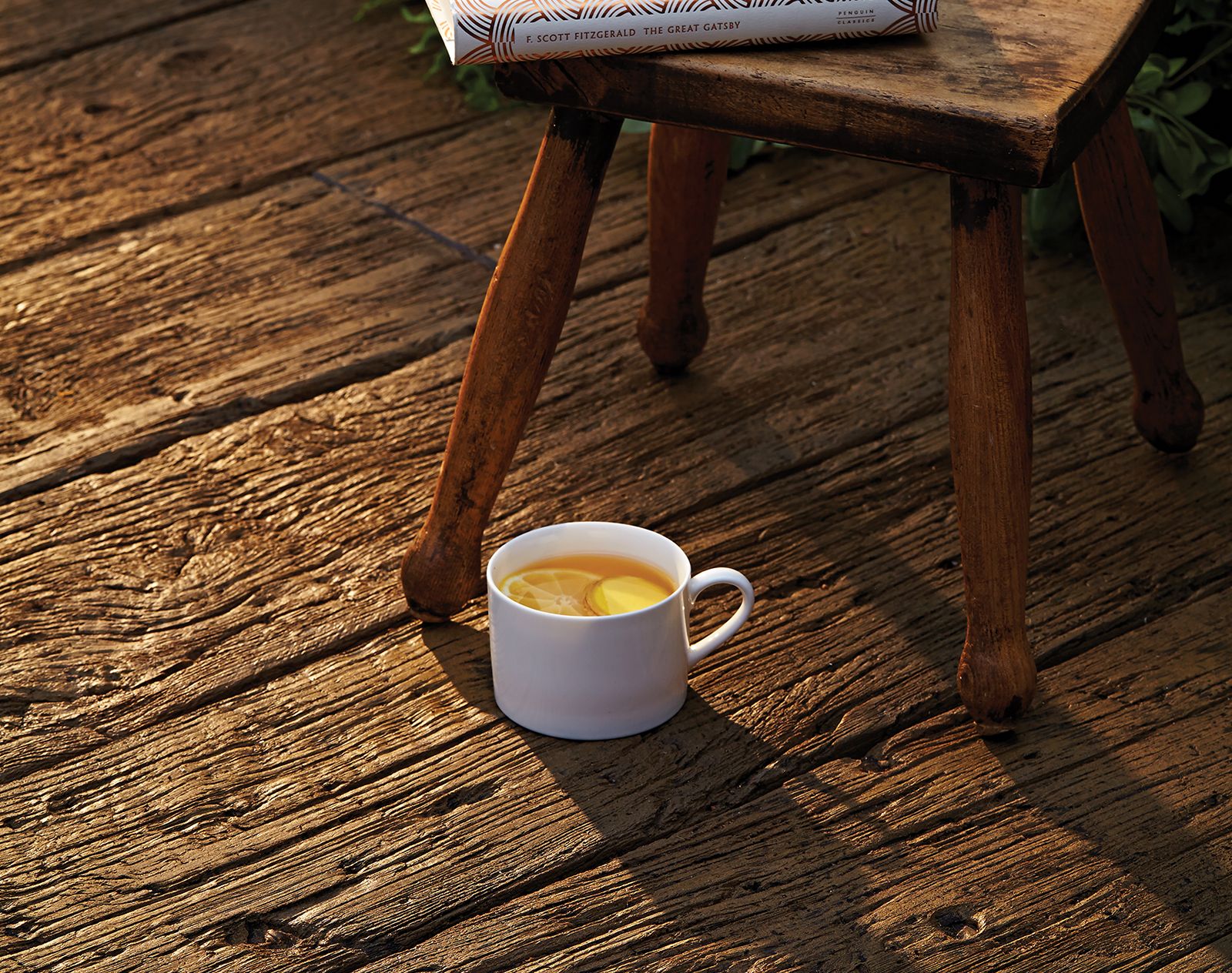 Rustic-style Millboard decking with enhanced woodgrain texture, featuring a white cup of lemon tea beside a wooden stool and a copy of The Great Gatsby.