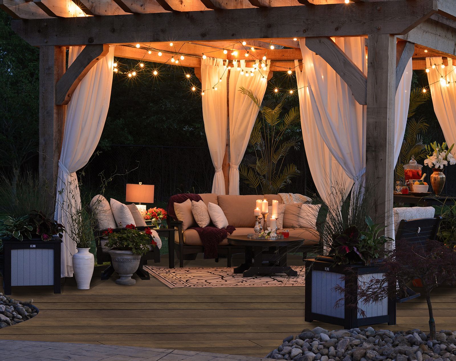 Millboard decking beneath a cosy outdoor pergola with draped curtains, ambient string lights, and cushioned seating, creating a stylish and inviting garden lounge area.