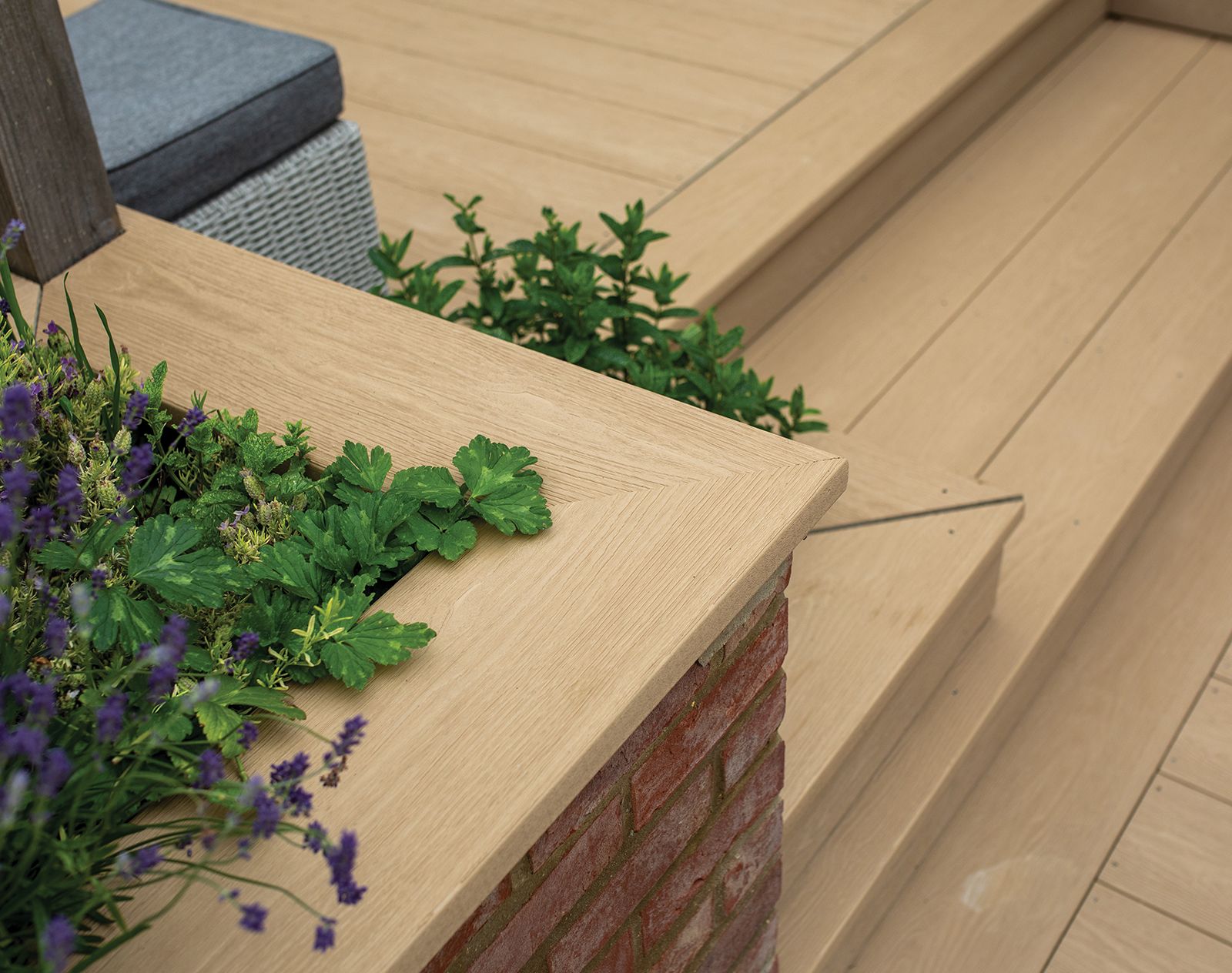 Corner detail of Nordeck PVC decking with integrated planter, steps and light wood-effect finish, surrounded by lush green plants and purple flowers.
