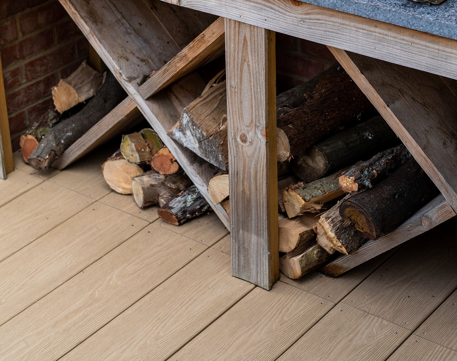 Natural wood-effect Nordeck PVC decking with stacked firewood stored in a rustic log rack, showcasing durable outdoor flooring.