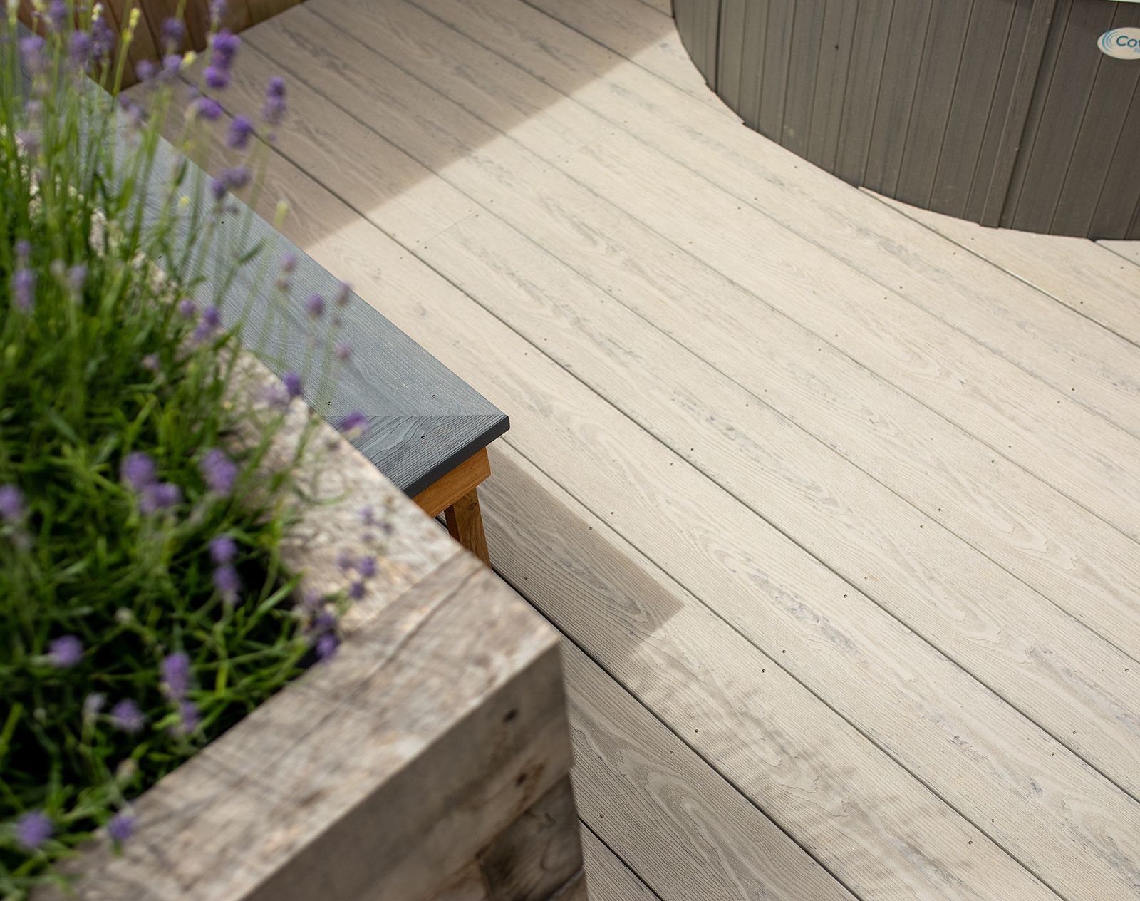 Close-up of light grey Nordeck PVC decking boards beside a hot tub and lavender planter, showcasing realistic wood grain finish. 