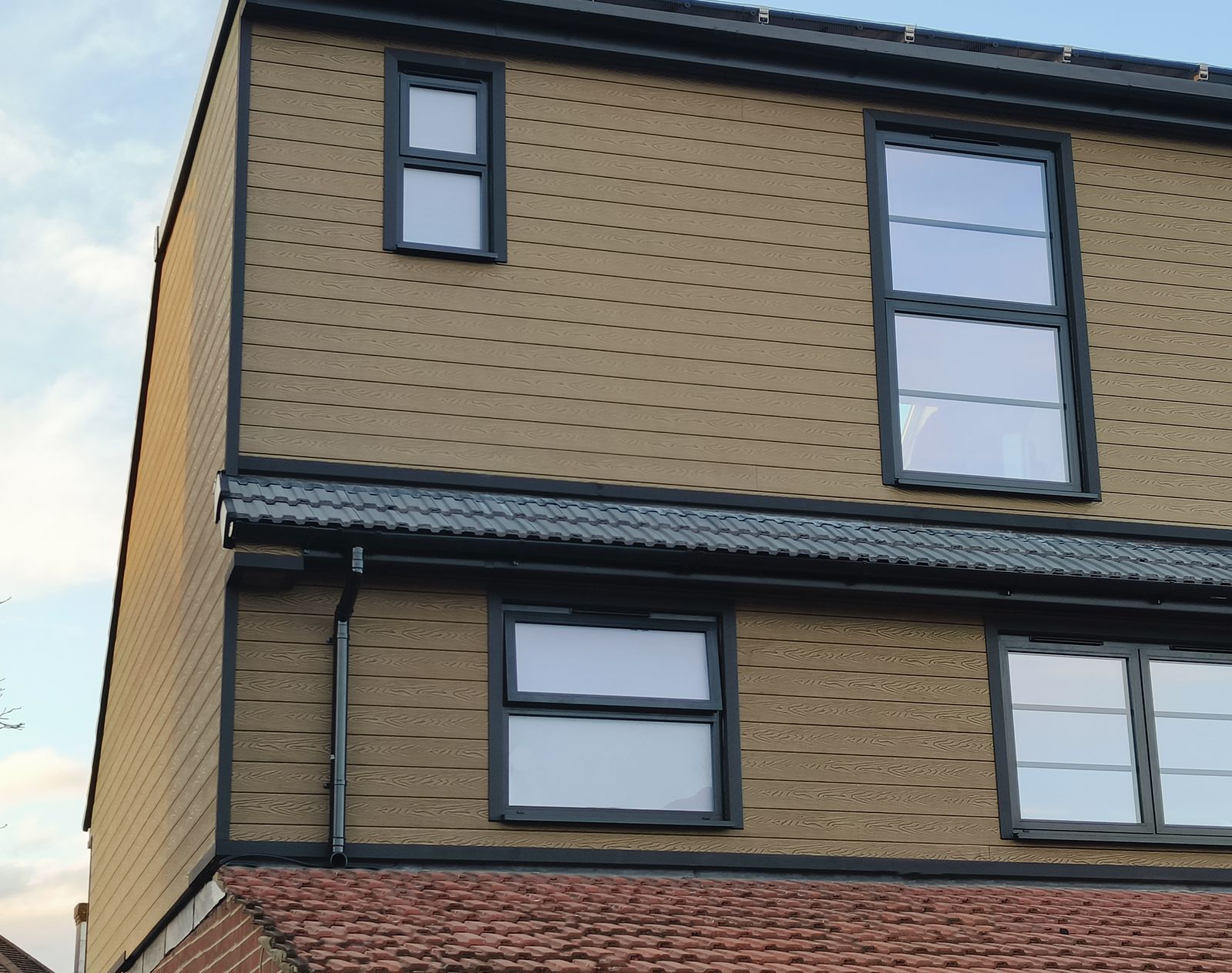 Modern house exterior finished with woodgrain-effect composite cladding in a natural oak tone, featuring black-framed windows and a tiled roof.