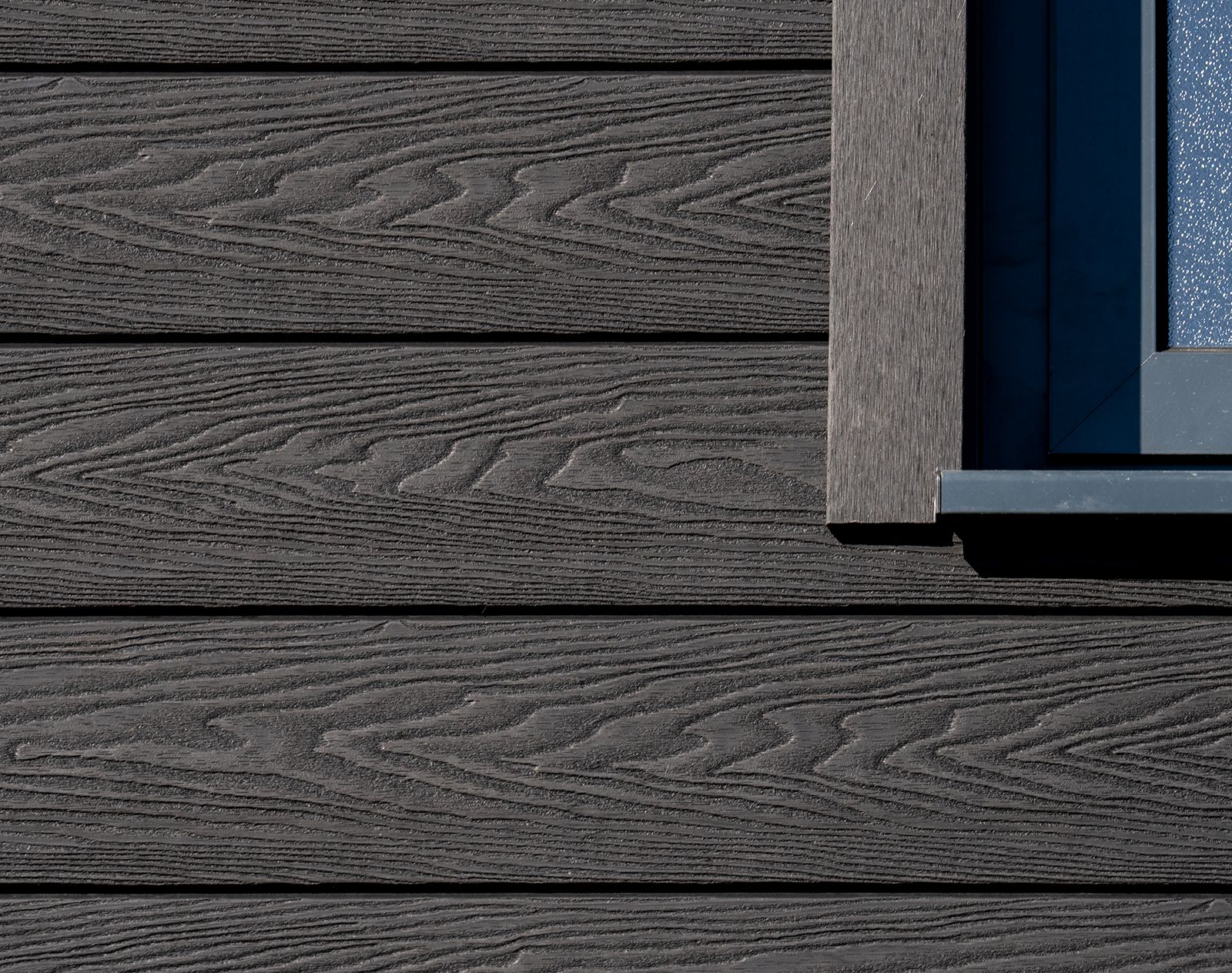 Close-up of black woodgrain-effect composite cladding boards with horizontal installation and matching trim around a modern window frame.