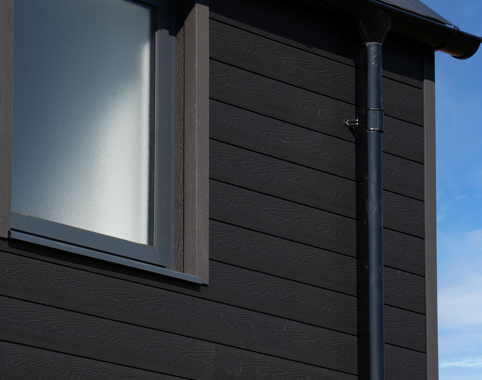 Side view of a house with black horizontal composite cladding, frosted window, and black downpipe, showing clean, low-maintenance exterior finish.