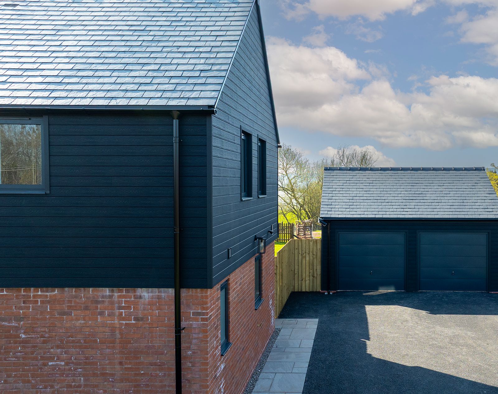Contemporary house and garage with black horizontal composite cladding on the upper walls and red brick on the lower level, finished with slate-effect roofing.