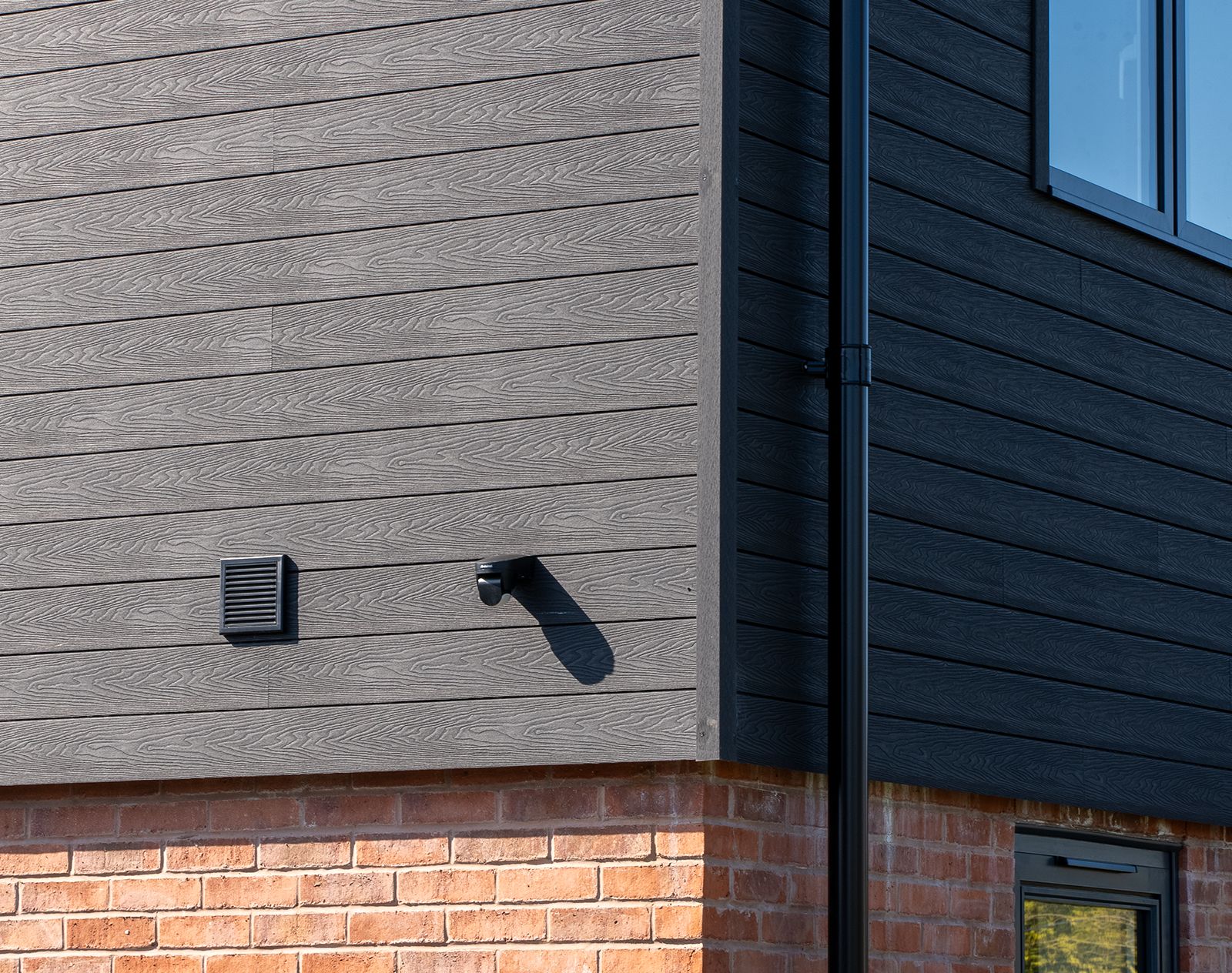 Corner detail of a modern home with dark grey composite cladding featuring woodgrain texture, installed above a red brick base with ventilation grille and security camera.