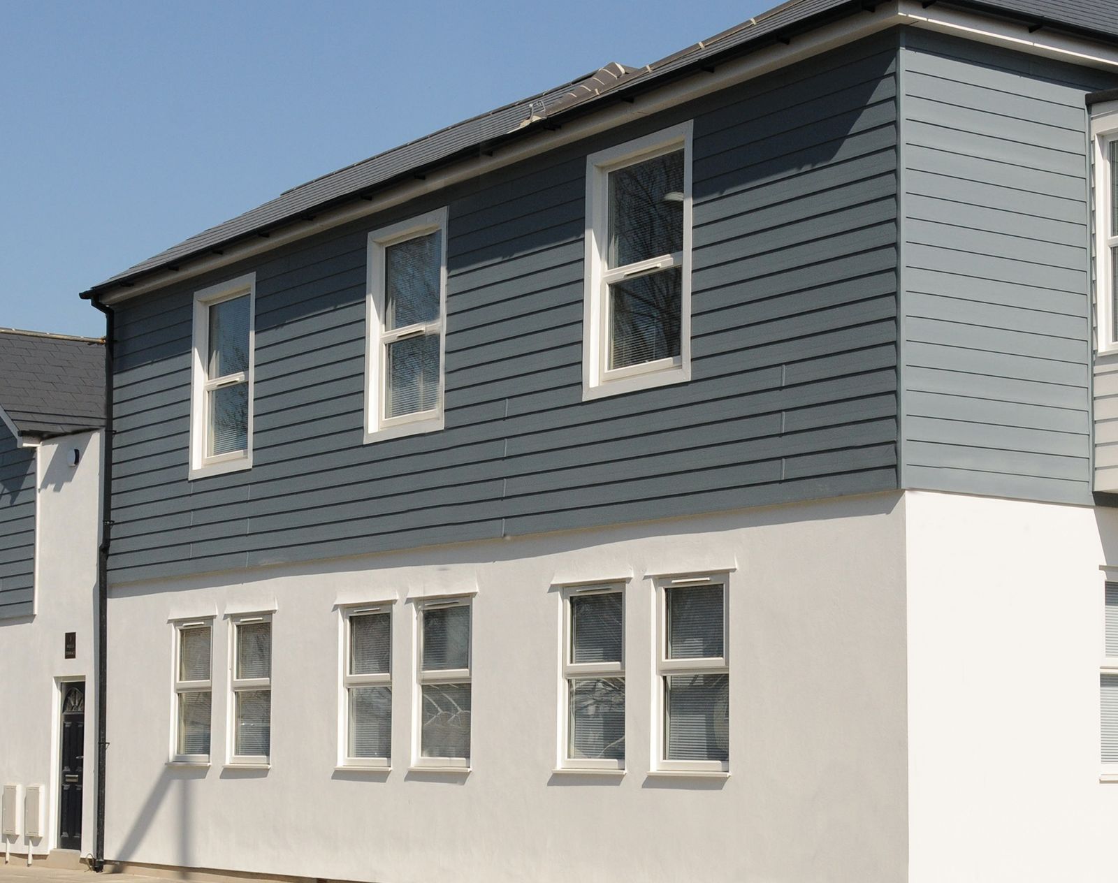 Modern residential building with grey horizontal composite cladding on the upper storey and smooth white render on the ground floor.