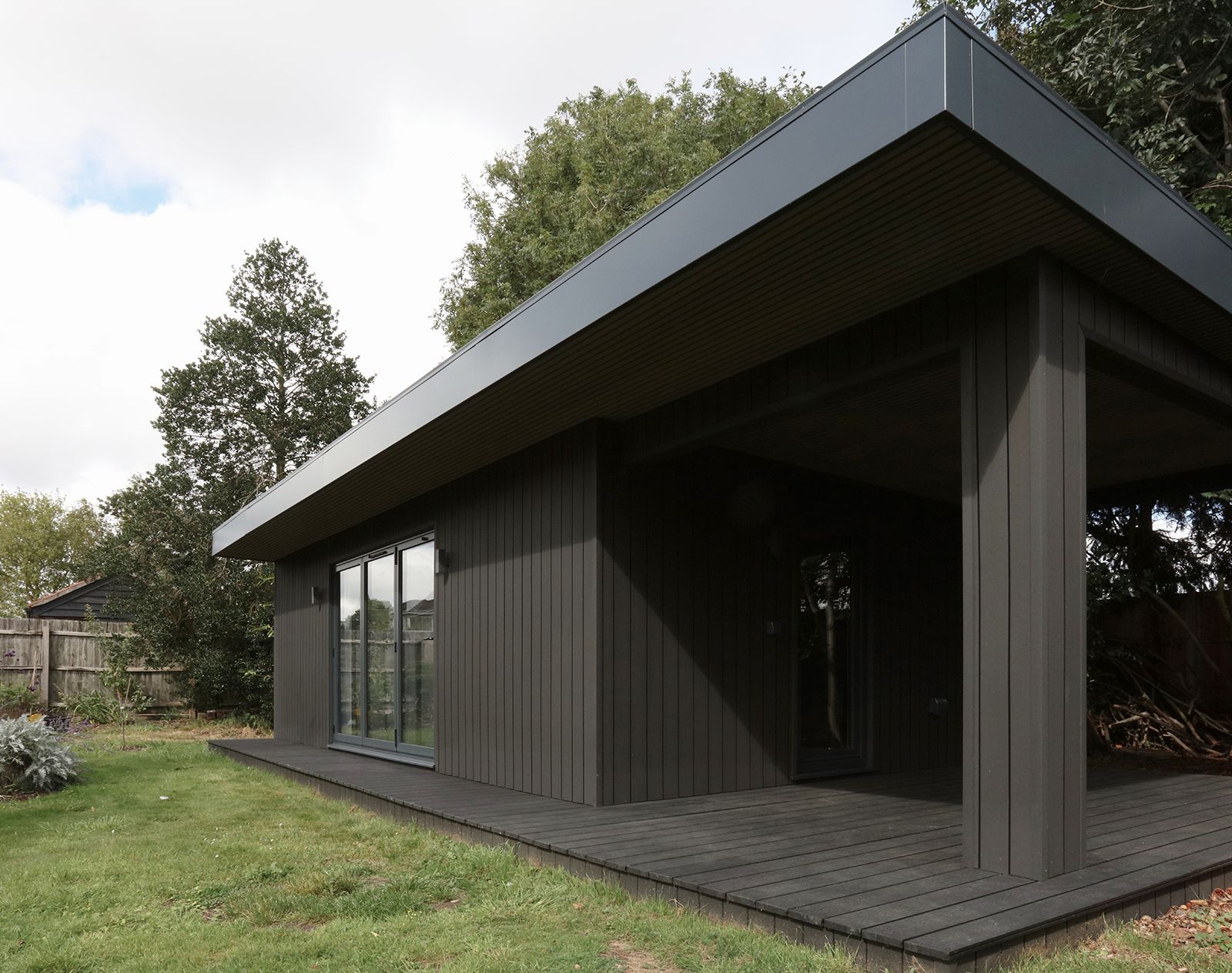 Modern garden building finished with vertical composite cladding in dark grey, featuring a flat roof, sliding doors, and wraparound decking.