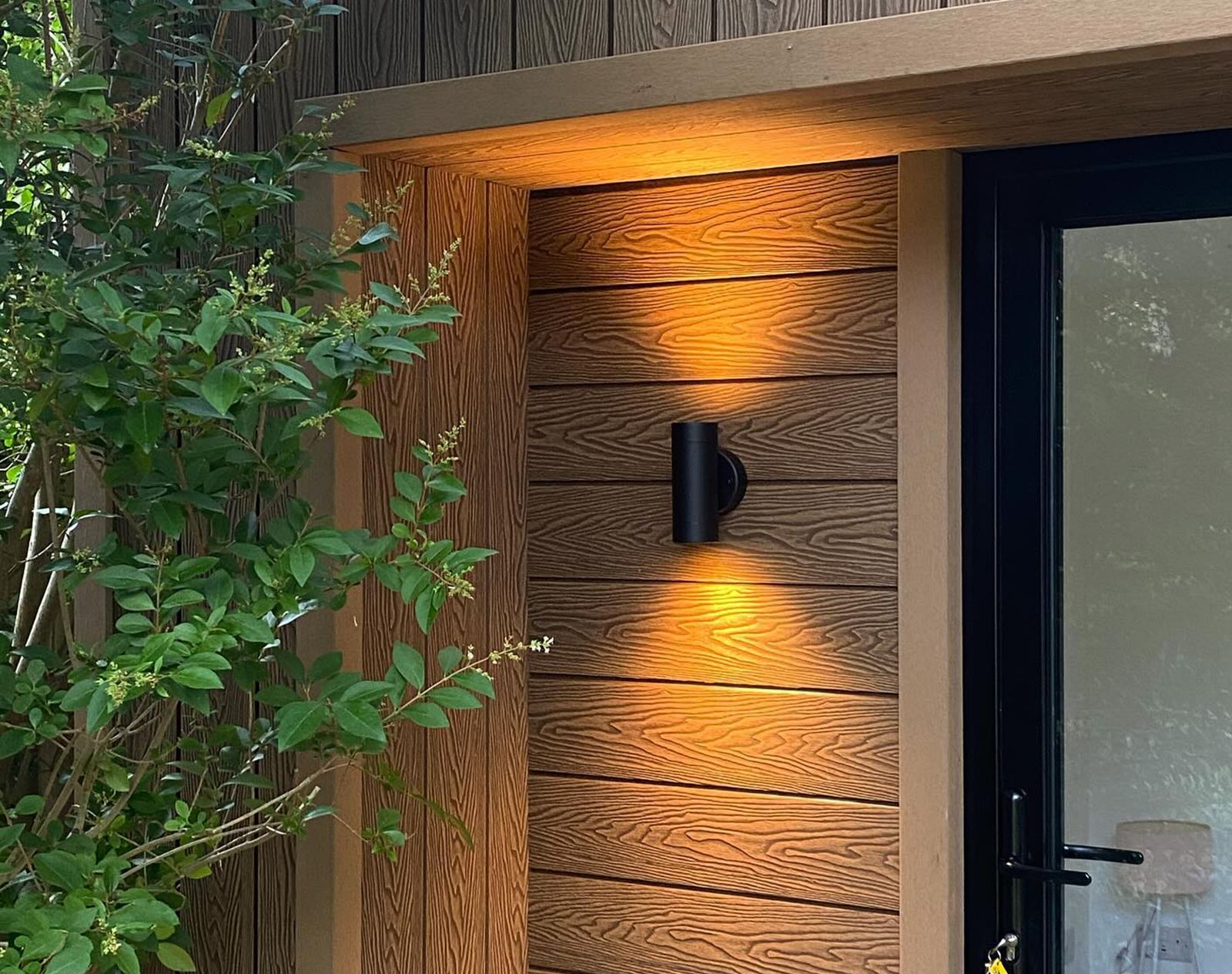 Natural wood-look composite cladding with warm grain texture, installed horizontally around a doorway with black outdoor wall light and glazed door.