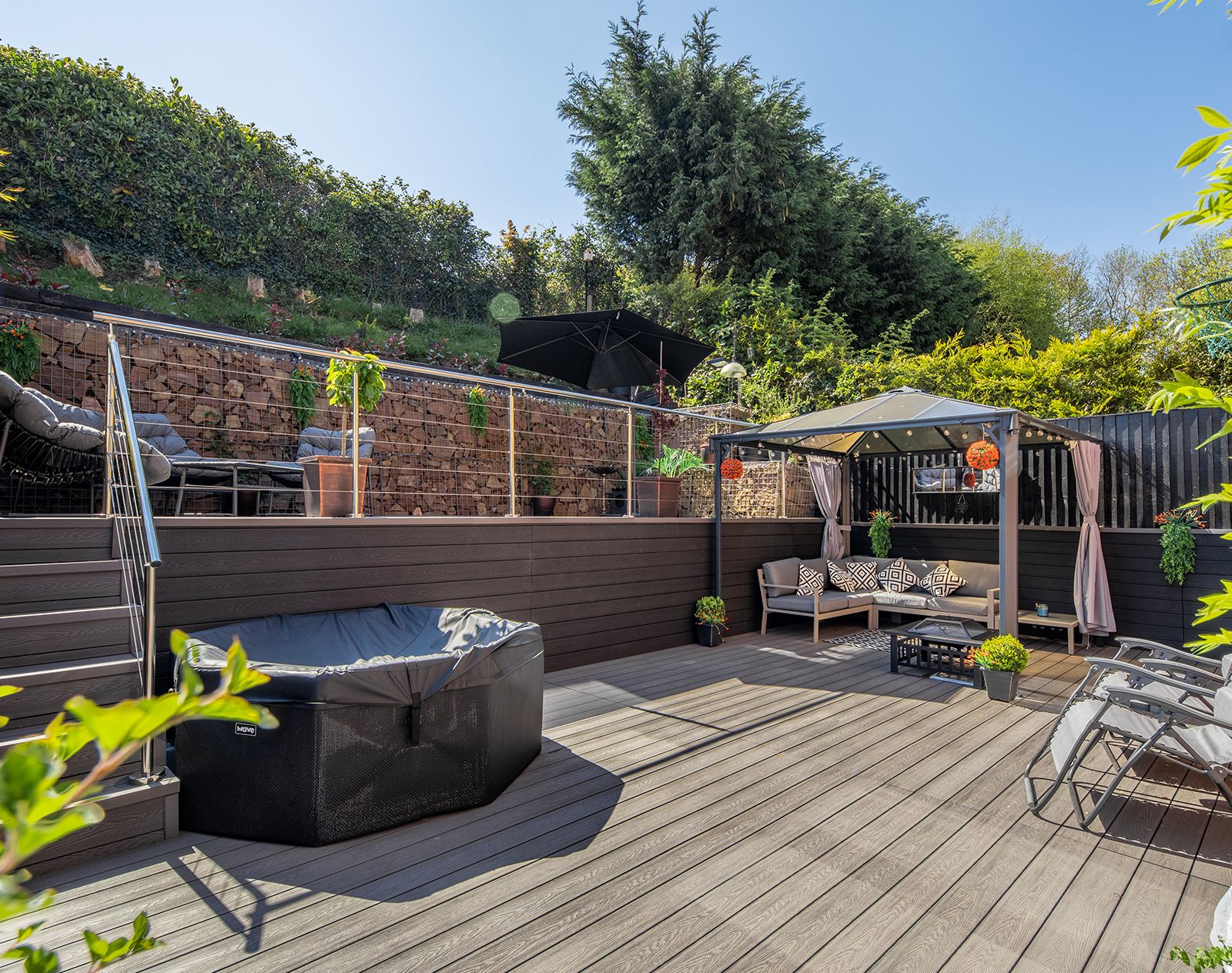 Two-tier garden with stylish grey composite decking, outdoor seating areas, pergola, and stainless steel balustrade, surrounded by lush greenery and decorative gabion wall.