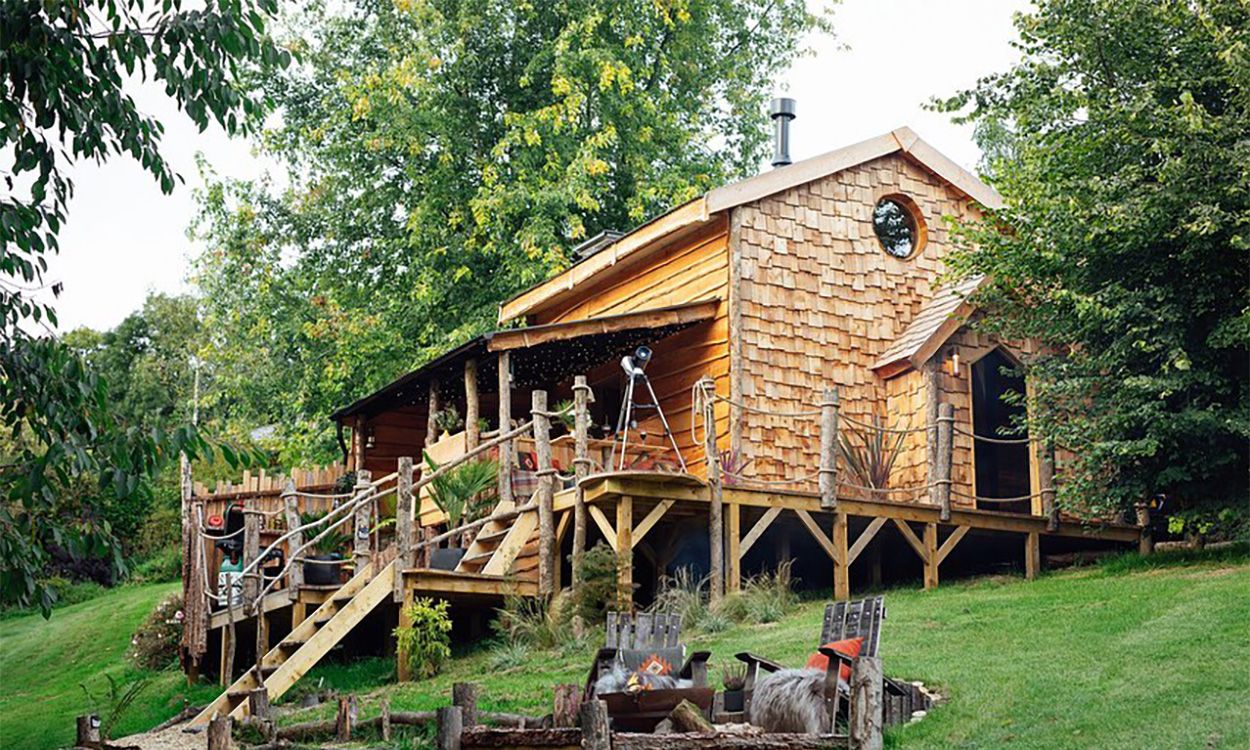 Ursa Bear Cabin handcrafted by With Bear Hands UK, featuring rustic timber shingles, Cladco Black corrugated steel roofing, and raised deck with woodland fire pit in Devon